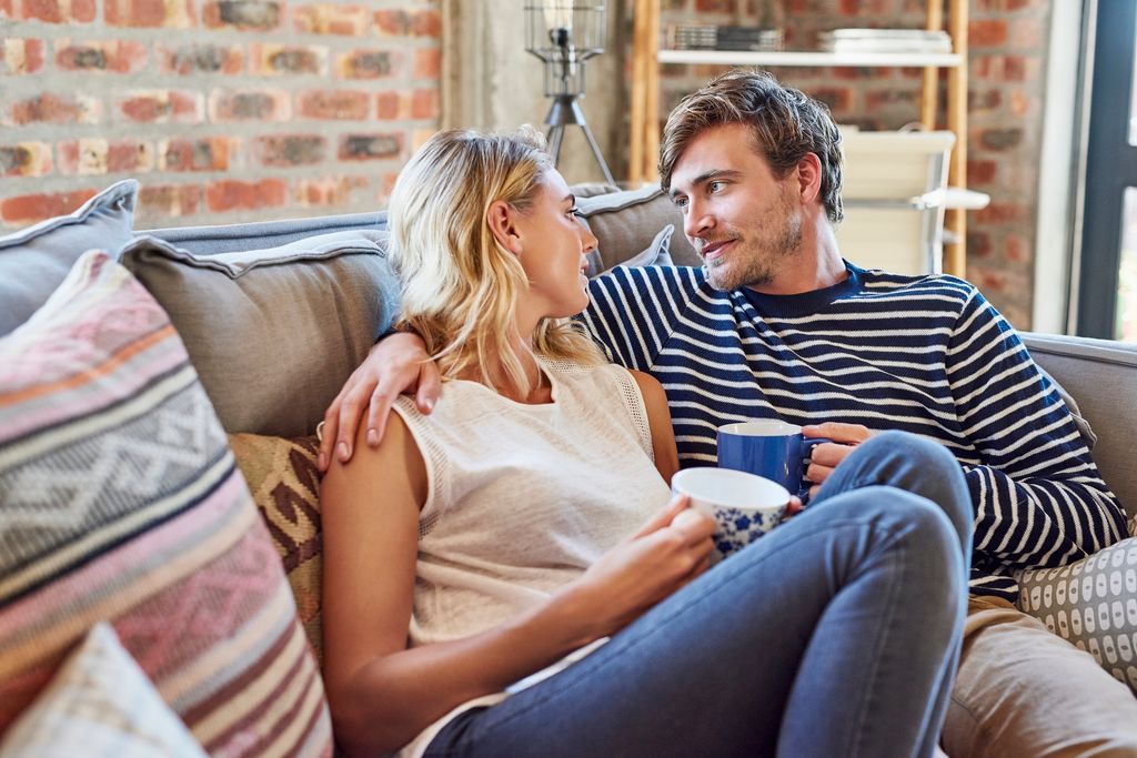 Romantic young couple looking at each other while relaxing on sofa. Man and woman are holding coffee mugs in living room. They are in casuals at home.