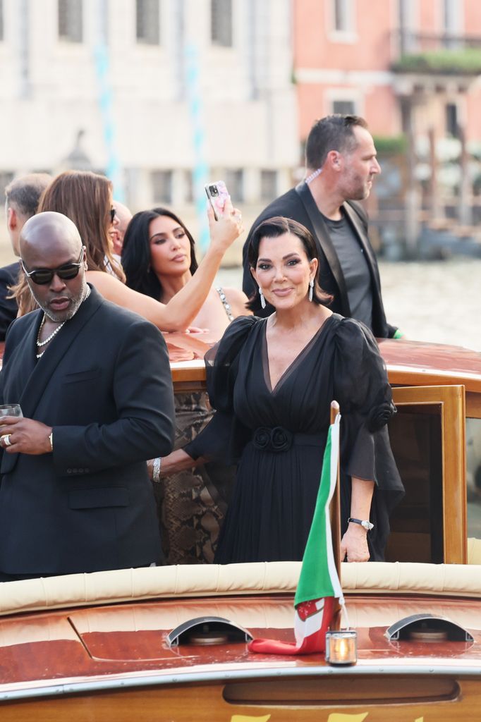 Kris Jenner se tient sur un bateau à Venise avec une longue robe noire avec des manches d'organza et une ceinture florale, accessoirisée avec des boucles d'oreilles et des cheveux courts élégants.