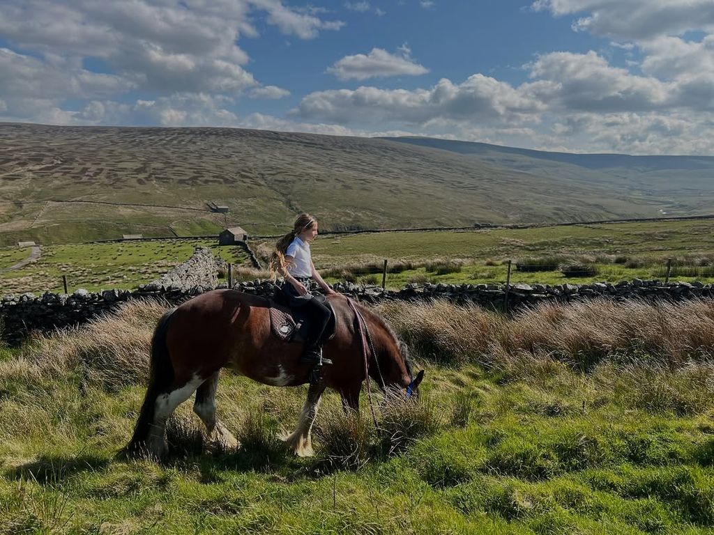 Amanda Owen's daughter sitting on a horse in a field in front of beautiful landscape 