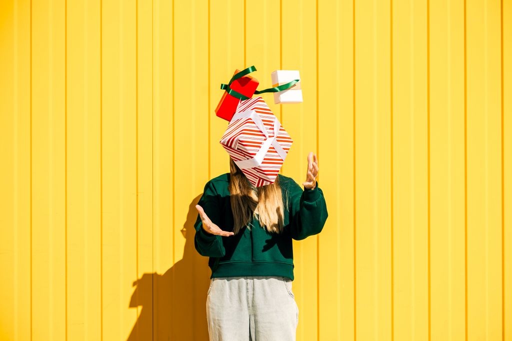 Woman in Christmas hat and green sweater catching Christmas gift boxes against the yellow wall. Funny summer Christmas and sales concept.