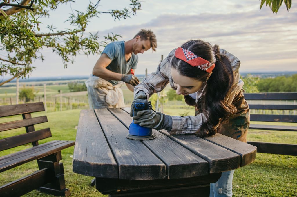 Adult couple,man with short brown hair and beard,woman with long brown hair,wearing apron,sanding their wooden table in the garden,woman using blue sanding machine in the forefront,face close to the machine,while her husband painting the places that have already been sanded in the background,serious focused facial expression,low angle,home improvement,horizontal