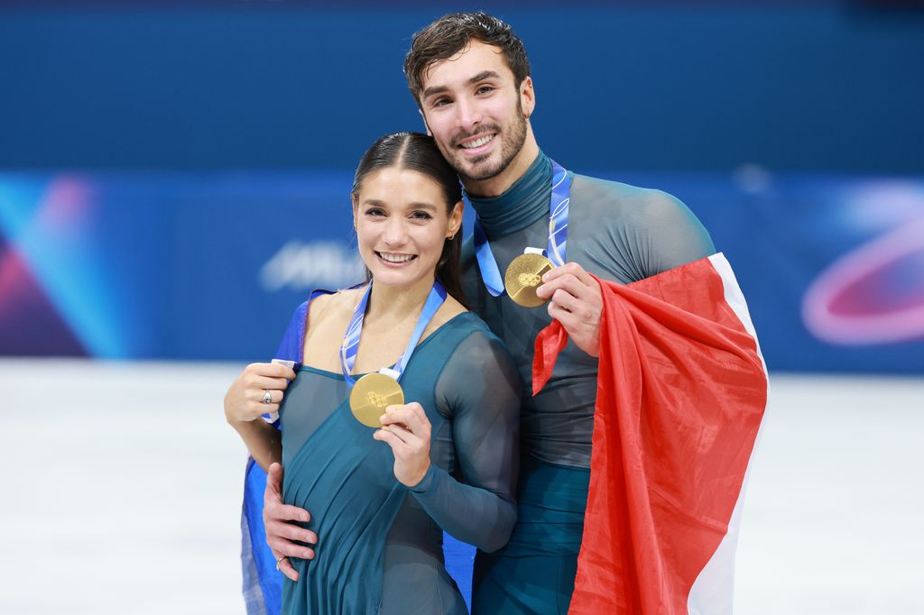Laurence Fournier Beaudry et Guillaume Cizeron tenant des médailles d'or sur la glace souriants, portant le drapeau français et des uniformes de patinage sur glace en maille bleue assortis