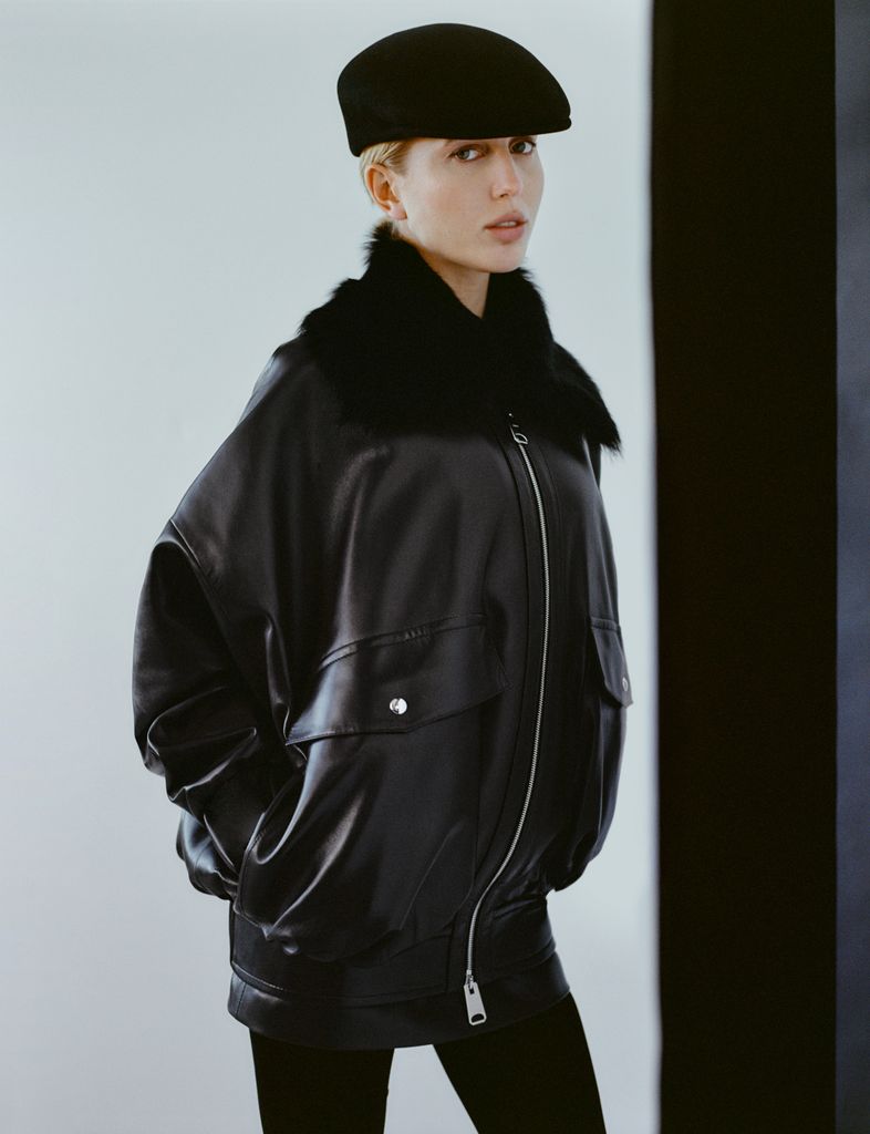 Princess Olympia of Greece poses in an oversized black leather bomber jacket with a faux fur collar, styled with black leggings and a sleek black cap. She gazes at the camera with a poised expression, while soft lighting enhances the moody elegance of the look.