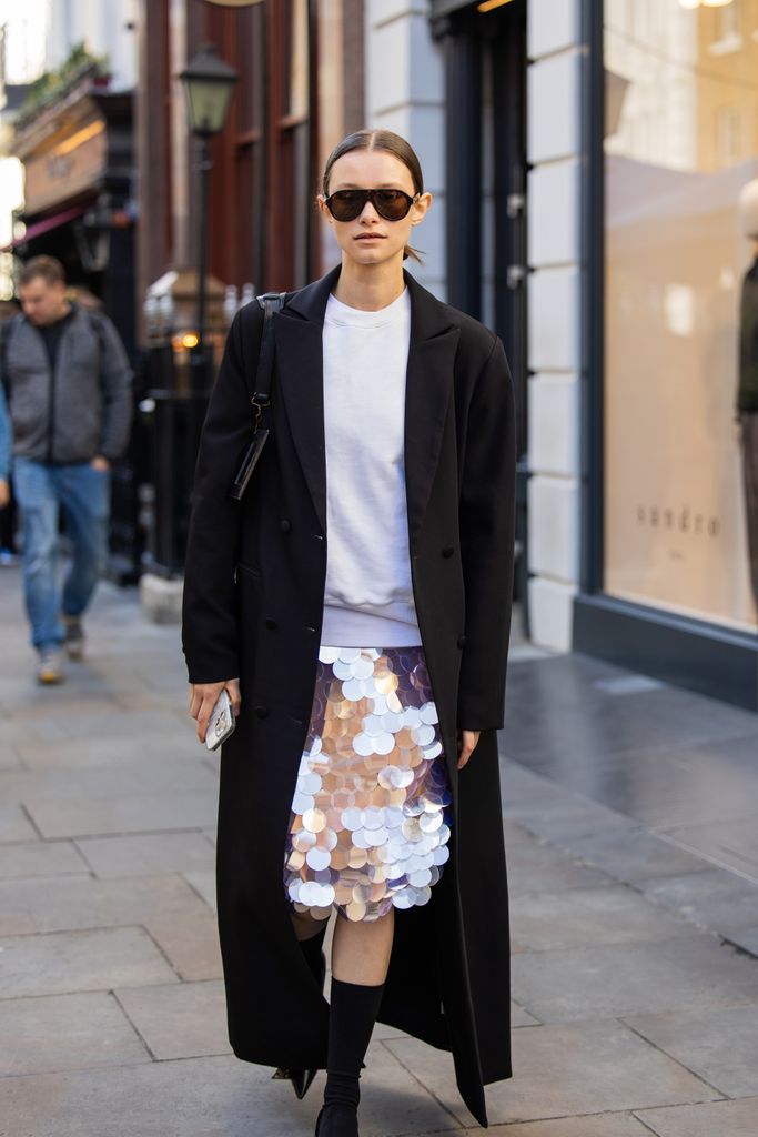 Ein Gast trägt einen Paillettenrock, einen schwarzen langen Mantel, eine Balenciaga-Tasche, eine Sonnenbrille und mittelhohe Socken vor Bora Aksu während der London Fashion Week im September 2024 am 13. September 2024 in London, England.