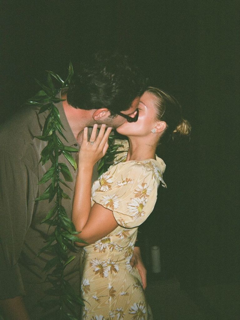 Sofia Grainge and her husband Elliot share a kiss, Elliot wears a green shirt and wears a garland of leaves around his neck, Sofia wears a yellow floral dress and her hair in a bun