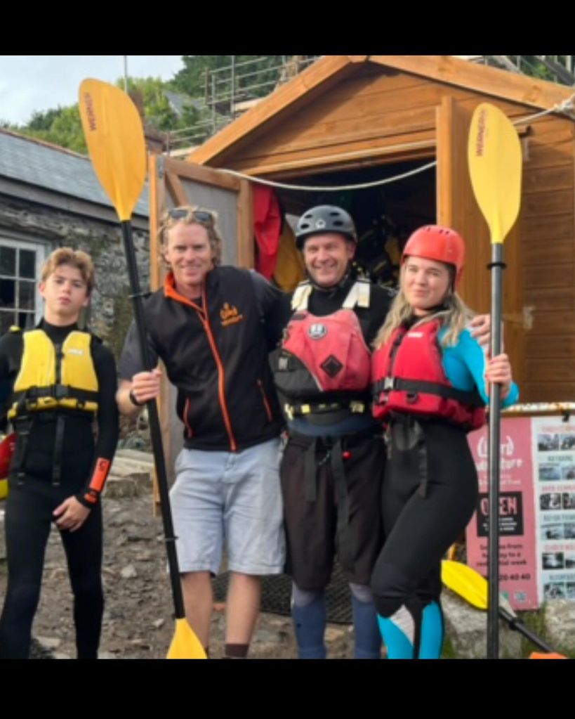 A photo of four people standing outside a shed, three of them wearing wetsuits 