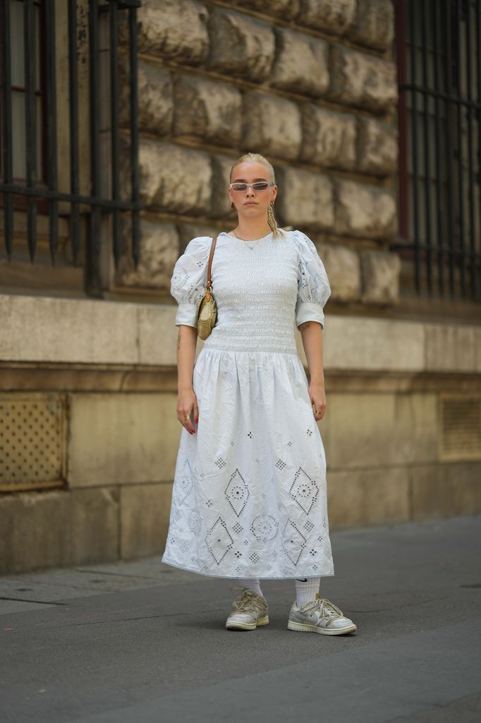 A guest wears silver sunglasses, gold earrings, a gold chain necklace, a white ruffled puffy short sleeves / long cut-out pattern dress, a beige denim with ripped GG logo monogram pattern shoulder bag from Gucci, white socks from Nike, white and pale gray matte leather sneakers from Nike x Off-White , outside Loewe, during the Menswear Spring/Summer 2024 