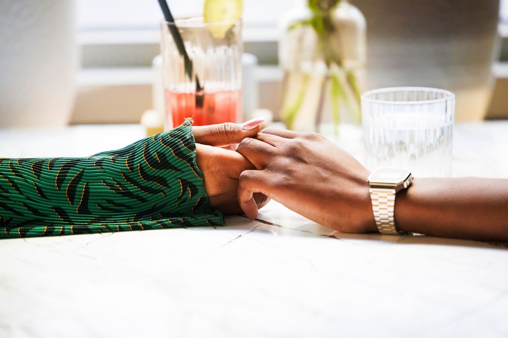 Two individuals hold hands across a table, conveying a sense of comfort and closeness in a relaxed setting.