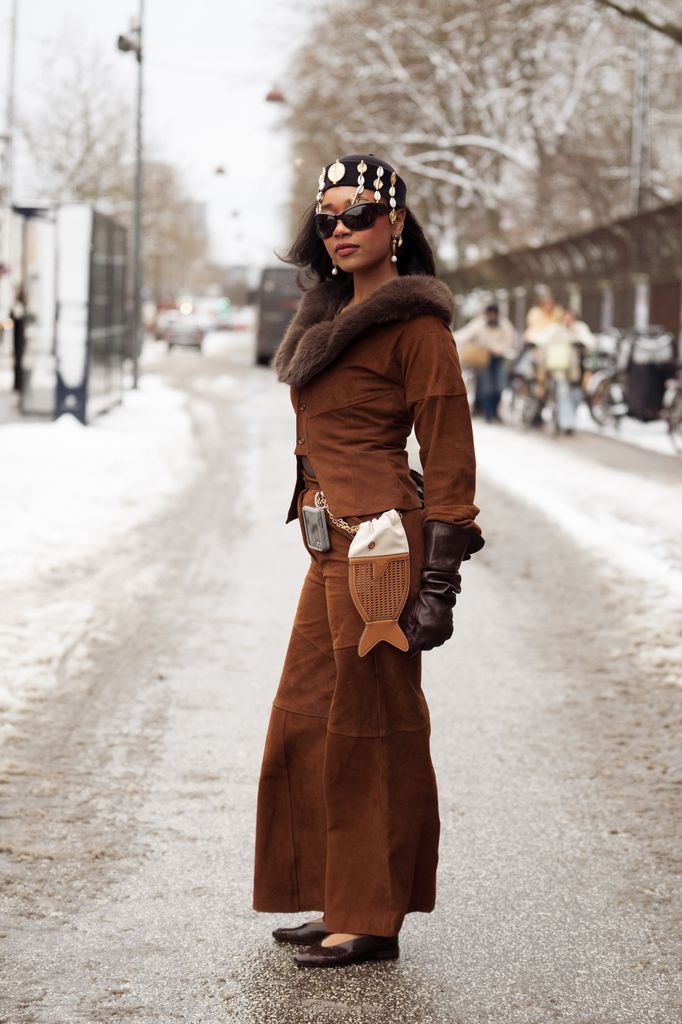 Petra Henriette Rufi wears a brown suede cropped jacket with a dark brown fur collar, matching high-waisted suede trousers, dark brown leather gloves, a decorative chain belt with a phone attachment, pointed brown heels, oversized black sunglasses, statement drop earrings, and a black headscarf embellished with shells and gold details outside the OperaSport show
