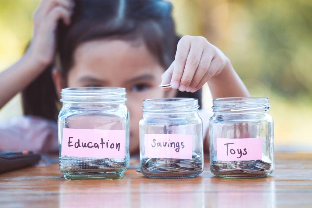 young girl putting money into glass jars marked education savings and toys