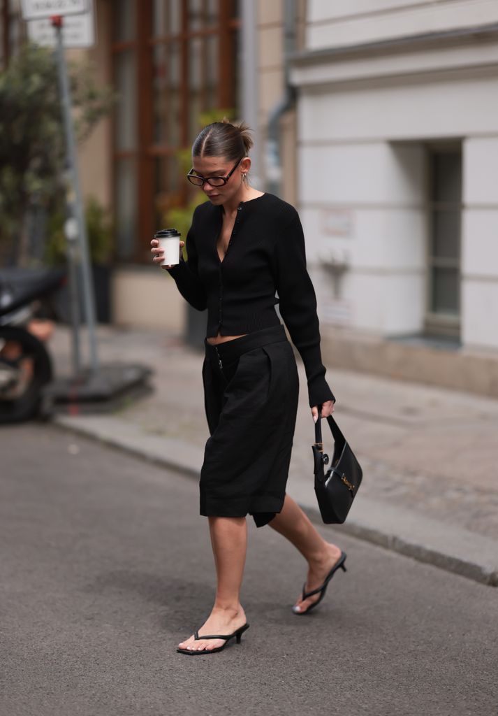 Sophia Geiss is seen wearing silver hoop earrings in two different sizes, oval brown glasses from Corlin, a silver nose ring, a black ribbed-knit cardigan with a fitted waistline and loose sleeves from & Other Stories, black pleated Bermuda shorts with wide leg from COS, a small rectangular black leather handbag with a gold YSL buckle from Yves Saint Laurent, and black kitten-heel sandals from LeGer, while holding a coffee cup