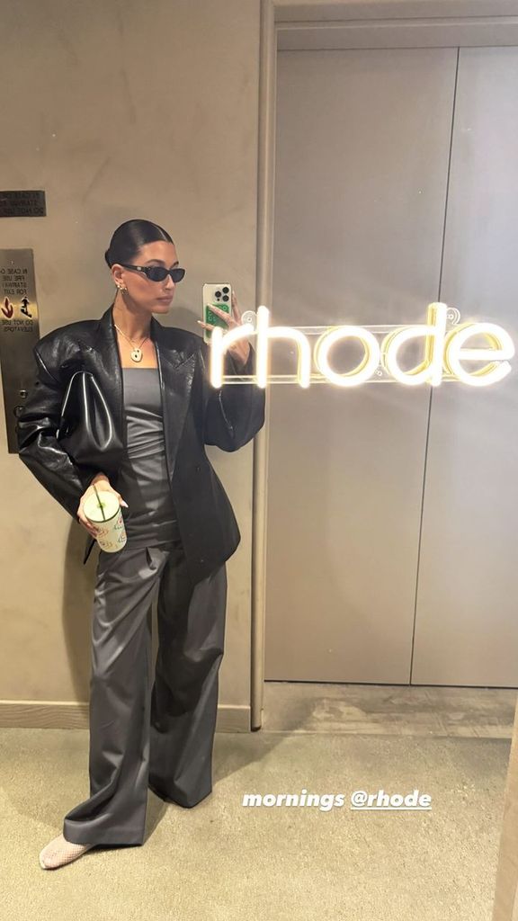 Hailey Bieber posts a mirror selfie at Rhode HQ wearing grey suit trousers and an oversized leather jacket