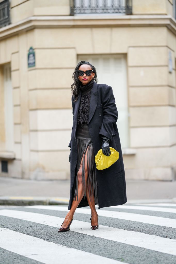 Emilie Joseph wears oversized double breasted wool extra long coat from COS, chocolate wool blended fringed turtleneck jumper from COS, midi length leather fringed Brown skirt with a thigh-high slit, lack leather long gloves, black sunglasses from Gentle Monster, a satin yellow clutch/pouch bag, burgundy / cherry / oxblood Lee Patent Leather pointed-toe stiletto/ slingback from Saint Laurent, Vintage looking square earrings, during a street style fashion photo session, on October 18, 2024 in Paris, France