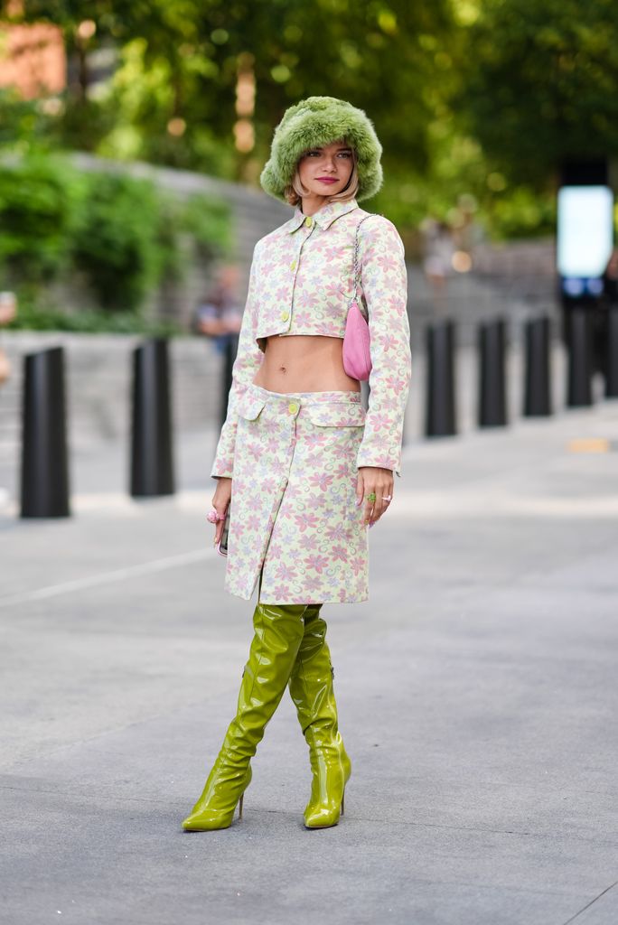 A fashion week guest wears a floral two piece, fluffy green hat and green latex thigh high boots