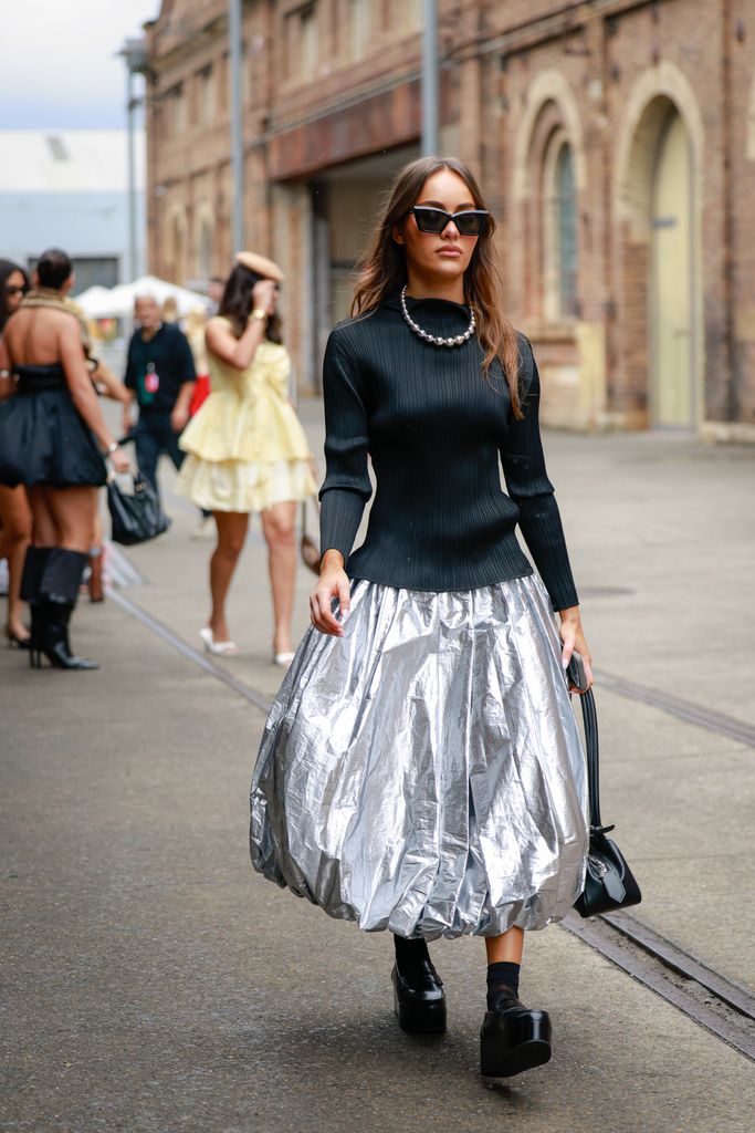 A guest wearing black top, silver necklace, black sunglasses, Comme des Garçons silver midi skirt and black loafers during Australian Fashion Week 2025 on May 15, 2025 in Sydney, Australia