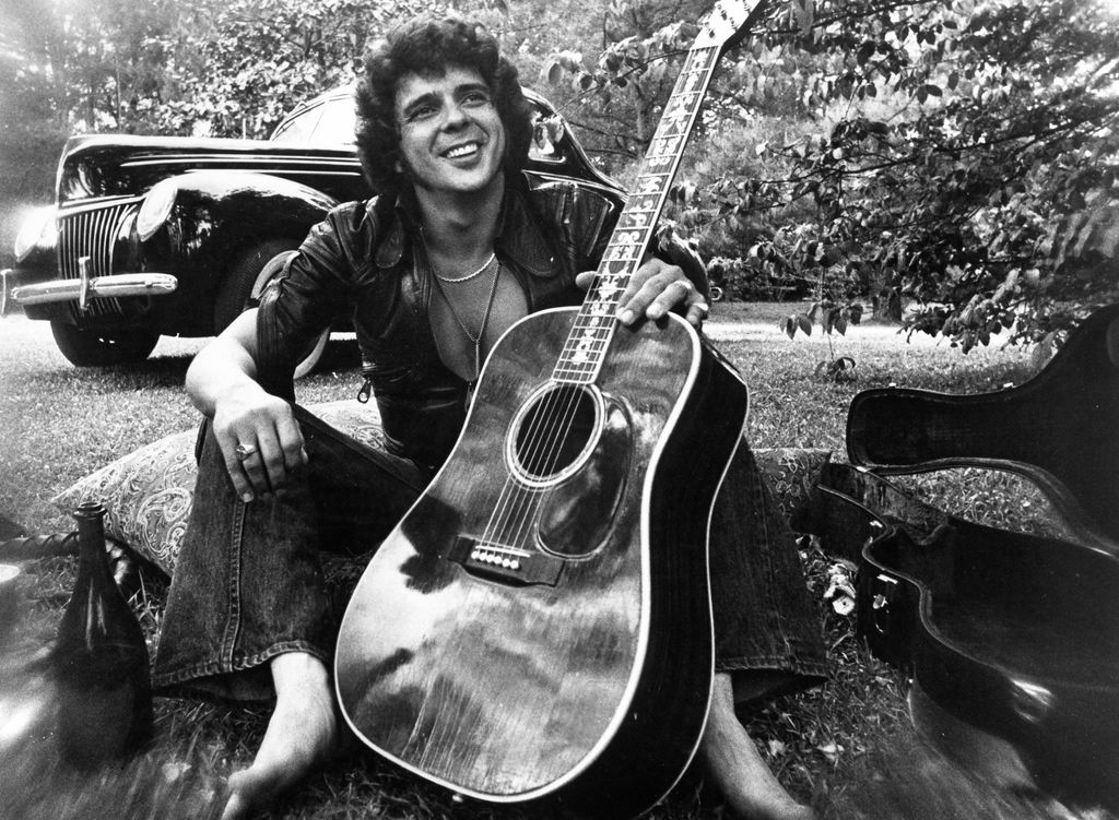 1975: Songwriter and guitarist Bobby Whitlock poses for a portrait in front of an old car next to a bottle of Dom Perignon champagne holding an acoustic guitar in circa 1975