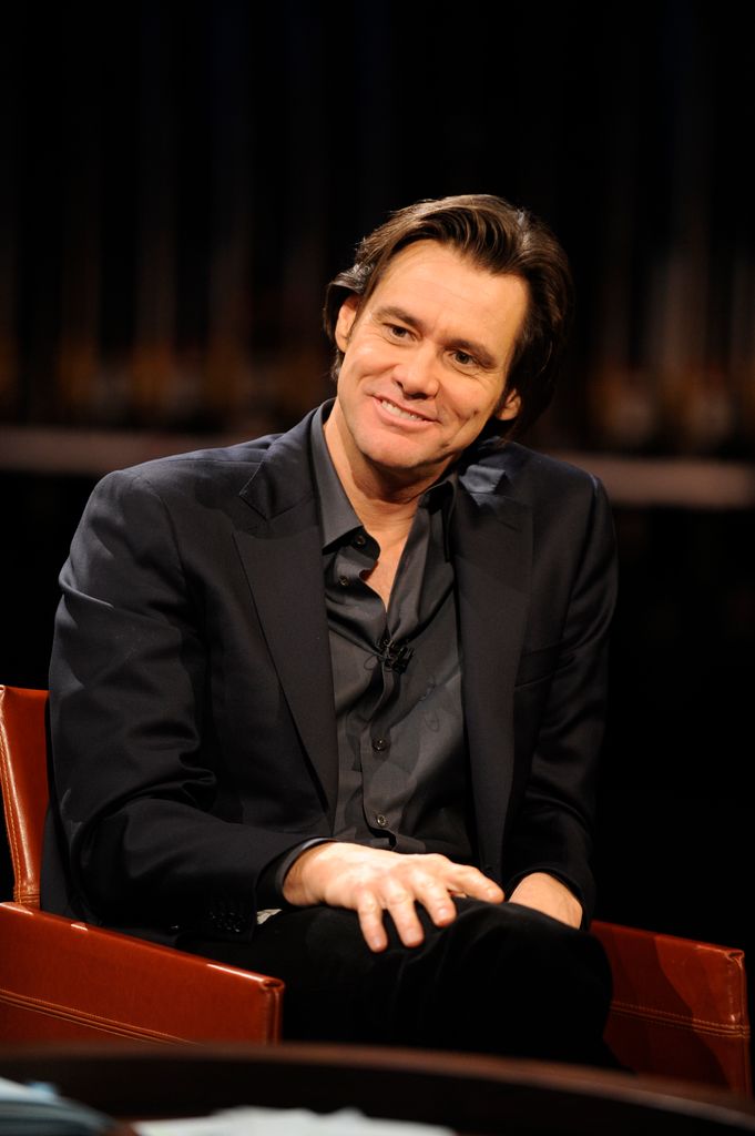 Jim Carey est assis sur une chaise rouge, les jambes croisées et tient ses mains sur ses genoux.