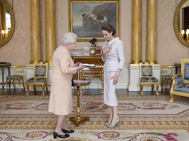 the queen stands in a beautiful reception room adorned with gold leaf furniture and a patterned gold and red rug wear a blush dress and greets a tall elegant woman wearing all white