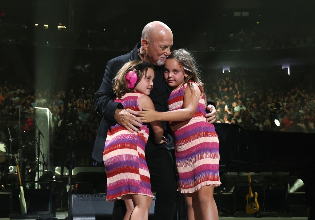 Billy Joel hugging his two daughters (Della Rose and Remy Joel) on stage, the girls are wearing matching pink patterned dresses