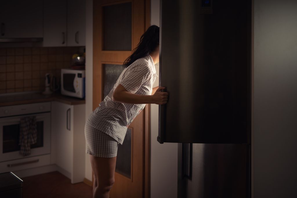 Woman leaning into fridge at night searching for food with bad diet habits.