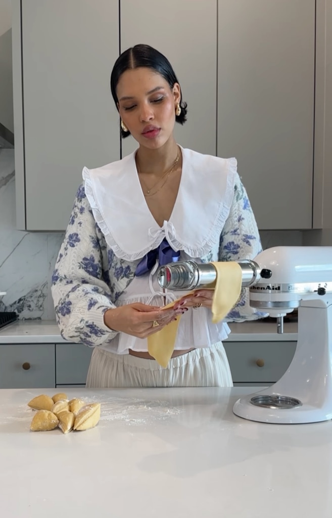 The image shows Nara Smith making pasta, operating a pasta machine. She wears a floral sweater with puffed sleeves, a large white collar, and soft, elegant makeup, looking focused and serene in a modern kitchen.