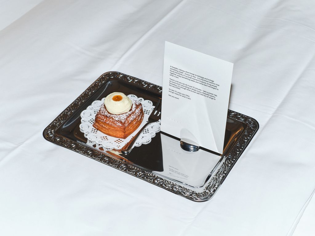 H! La mode rencontre le nouveau Volvo EX60 révolutionnaire 1 Une pâtisserie suédoise est posée sur un plateau d'argent sur un lit d'hôtel - un morceau de papier explique l'art du « Fika ». Le terme suédois pour faire une pause pour se ressourcer