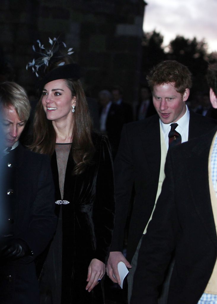 Kate Middleton in a plunging black dress and fascinator smiling next to Prince Harry