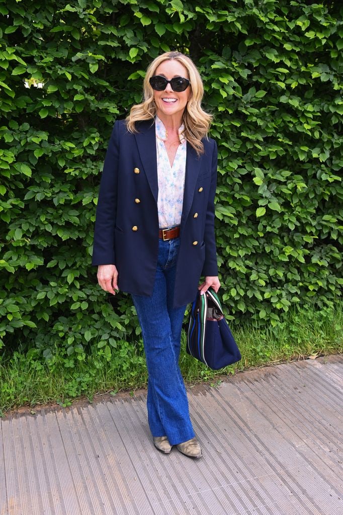 Jane Krakowski wears a navy blazer, floral shirt, flared jeans and sunglasses at the Chelsea Flower Show, standing in front of greenery.