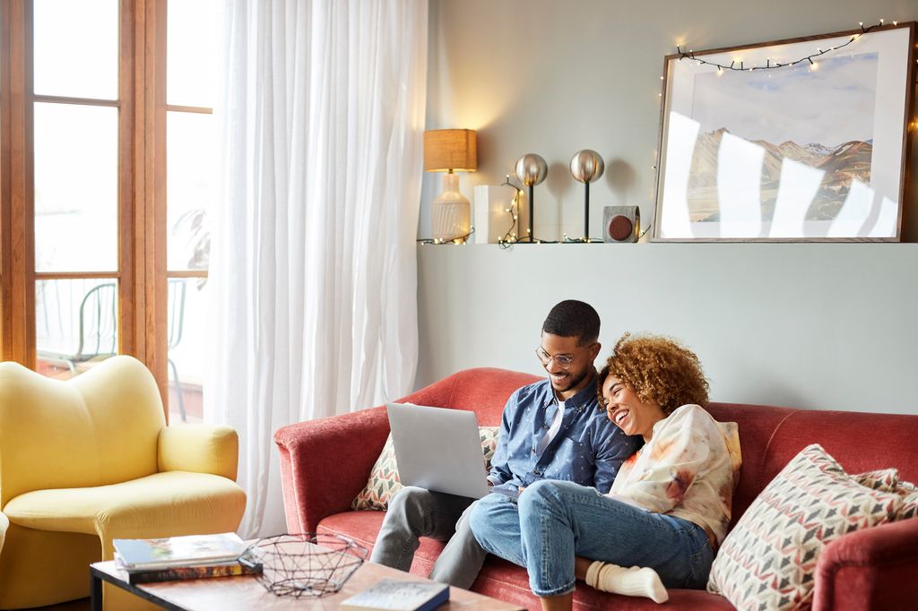Afro woman leaning on man using laptop in living room. Young couple is spending leisure time at home. They are sitting on sofa.