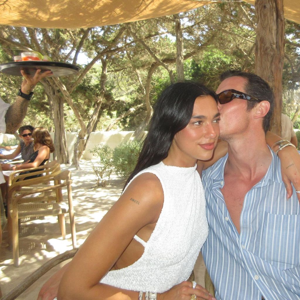 Callum Turner kisses Dua Lipa in a restaurant surrounded by trees. Callum wears a blue and white striped shirt with sunglasses