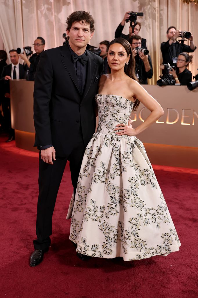 Mila Kunis and Ashton Kutcher on the red carpet, Ashton wears a dark suit and matching shirt and tie, while Mila wears a black and white floral strapless dress