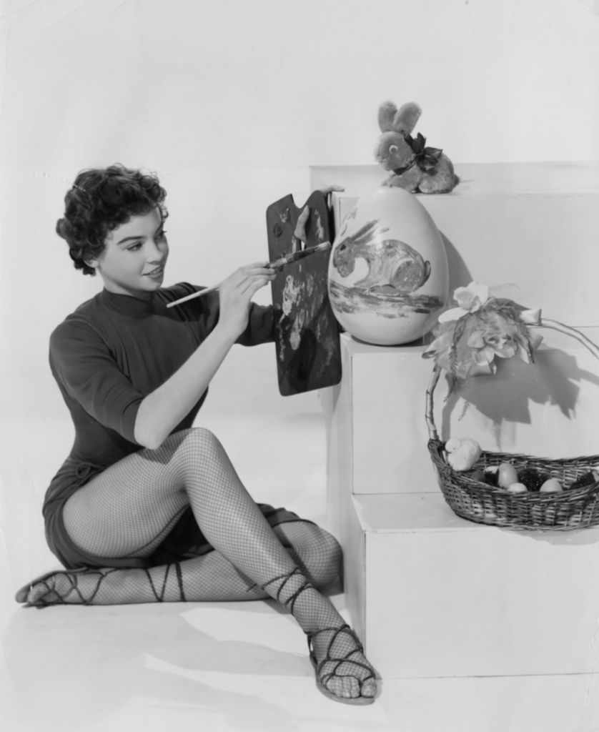 actress Leslie Caron in fishnet tights and heels paints a large Easter egg with a palette and brush.