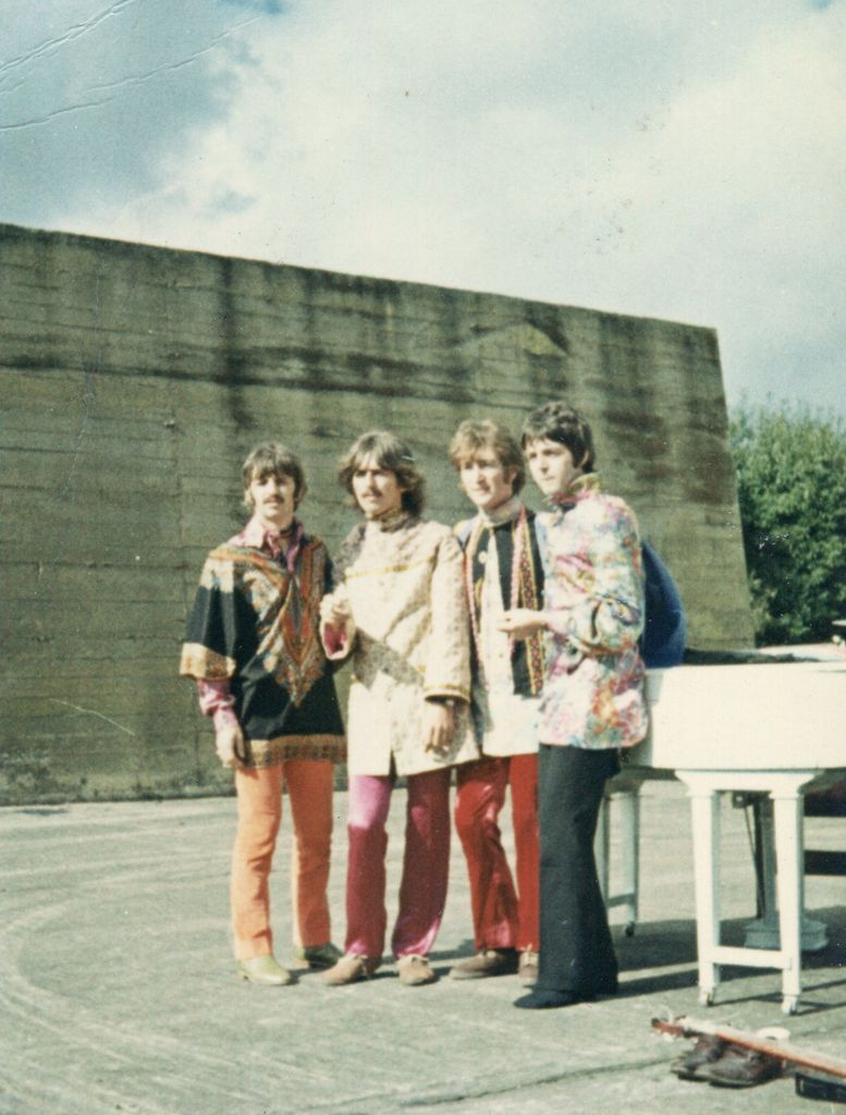 The Beatles wear ornate, colorful Eastern-inspired outfits with flared pants and paisley prints, standing next to a white grand piano outdoors. The sky’s blue, and so is the vibe.