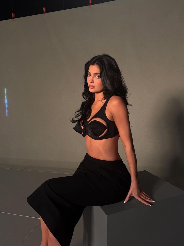 In a moody studio setting, Kylie Jenner sits confidently on the edge of a matte grey block against a neutral background. She wears a sculptural black bra top that dramatically contours and lifts, featuring a striking cutout design reminiscent of Madonna’s iconic cone bra era. Her voluminous dark hair cascades in soft waves, framing a glamorously contoured face with a nude-pink pout. A high-waisted black skirt hugs her curves and flows just below the knee. Her manicured nails are long, sharp, and white — a modern femme fatale finishing touch. The lighting creates subtle shadows, lending a cinematic quality to the image, as if Kylie has just stepped off a 90s runway or into a modern-day fashion noir.