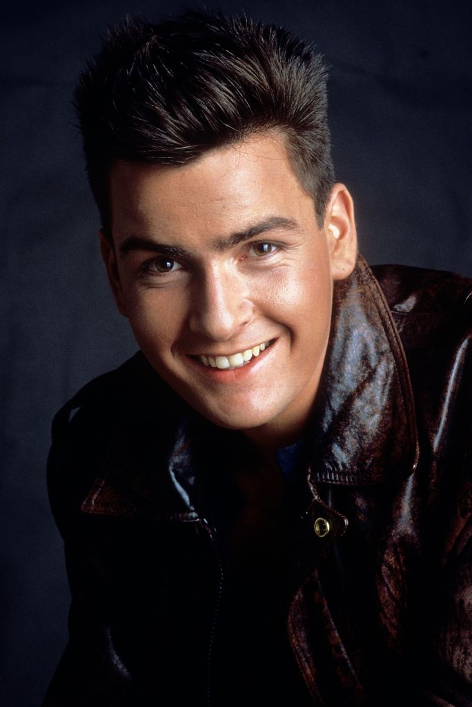 Charlie Sheen porte une veste en cuir et sourit largement à la caméra dans un portrait sombre.