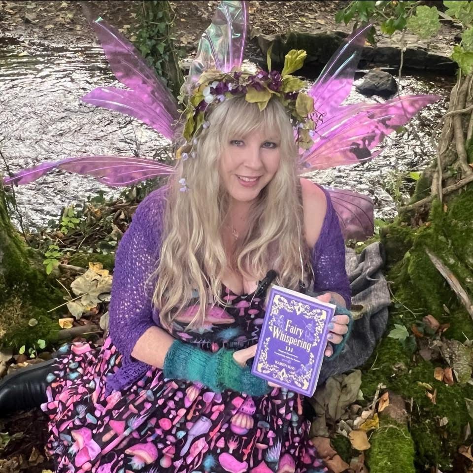 An image of Karen Kay, wearing fairy wings, and holding a copy of her book Fairy Whispering