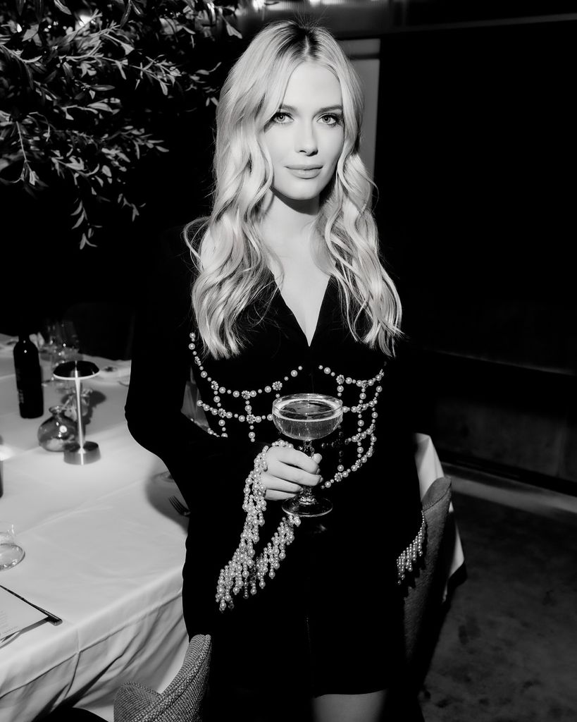 A black and white photo of Lady Eliza Spencer wearing a black mini dress and holding a glass of champagne. Her hair is down and wavy and she is looking directly into the camera