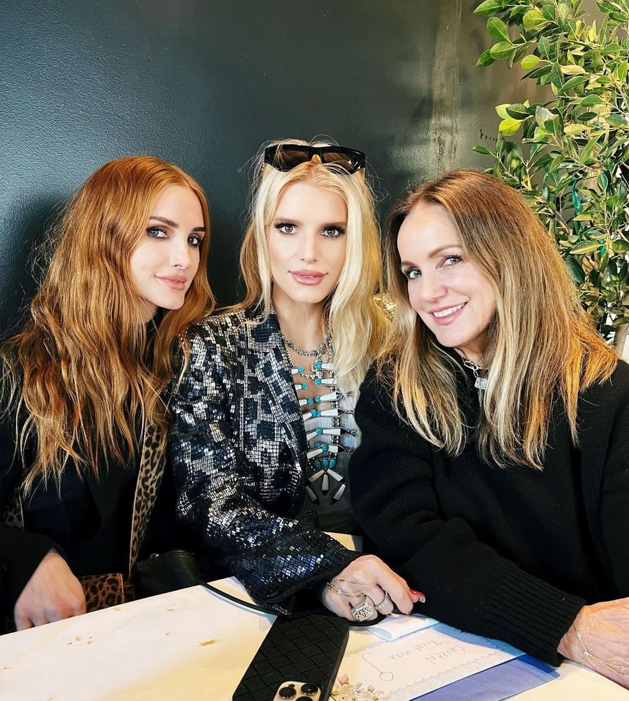 Ashlee Simpson and Jessica Simpson sit with their mom Tina at a table with a plant behind them