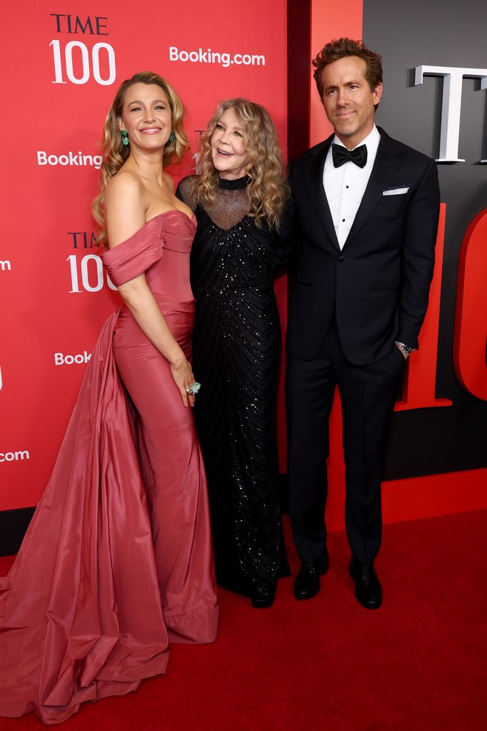 Blake Lively wears a red off the shoulder dress and stands next to her mum Elaine who wears a black sparkling dress and husband Ryan Reynolds who is wearing a black suit with white shirt and black bow tie 