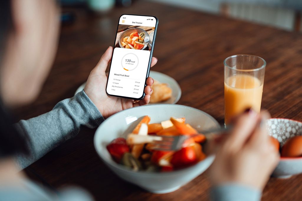 Over the shoulder shot of young woman using mobile app to track nutrition and count calories with smartphone while eating breakfast. Health eating lifestyles concept. Morning ritual.  Welcoming a brand new day.