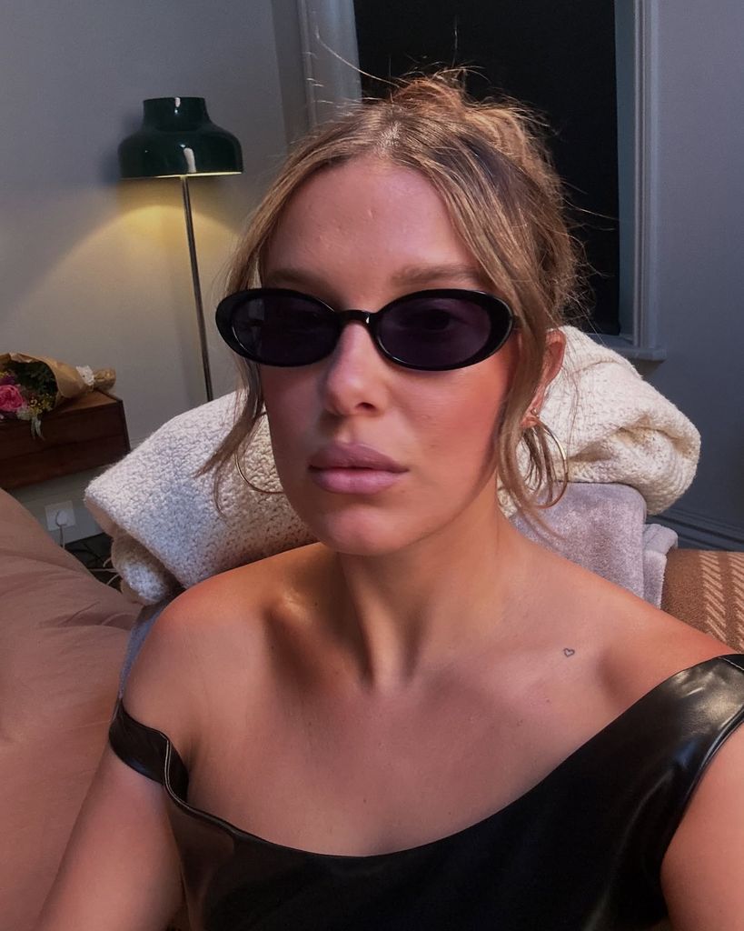 In this close-up selfie, Millie Bobby Brown is seated indoors, facing the camera with a pouty expression. She is wearing sleek, oval-shaped black sunglasses reminiscent of early 2000s fashion and a one-shoulder black leather-look top, which reveals minimalistic tattoos—a small outline of a heart near her collarbone and the Vivienne Westwood orb on her forearm. Her light brown hair is styled up with a few soft tendrils framing her face. Gold hoop earrings complete the Y2K-inspired look. The background includes a pink bedspread, a bouquet of flowers on a nightstand, and a gold-wrapped gift box, suggesting a celebratory or intimate setting. 