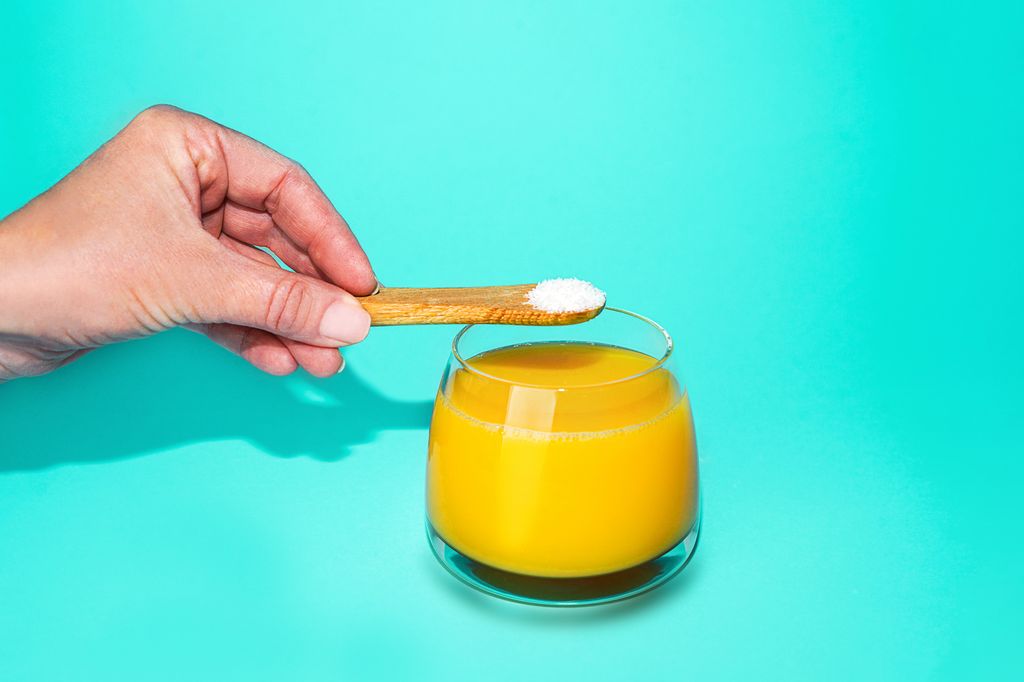 Hand with spoon of protein or collagen and orange juice on mint green color background. 