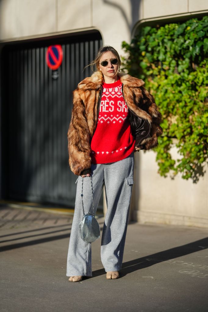 Emy Venturini wears black Rayban sunglasses, a red and white Yoox pullover, a brown faux fur jacket, grey wide-leg Yoox pants, a silver Rabanne bag, a light brown / cream leather Margiela shoes high heels, during a street style fashion photo session, on January 07, 2025 in Paris, France.