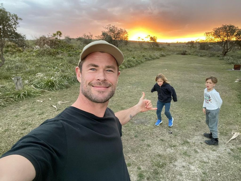 Chris Hemsworth smiling and taking a selfie with his two twin boys behind him playing in a field. The sun is setting behind them.