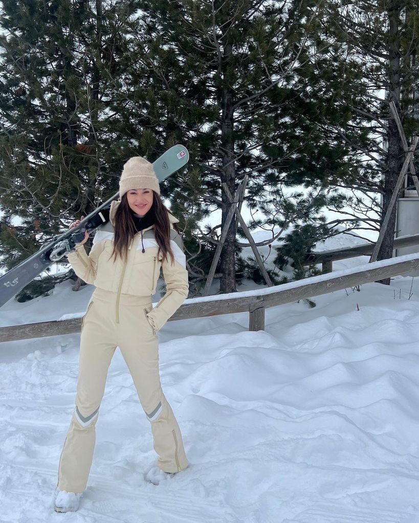 Michelle in a beige ski suit with skis on shoulders