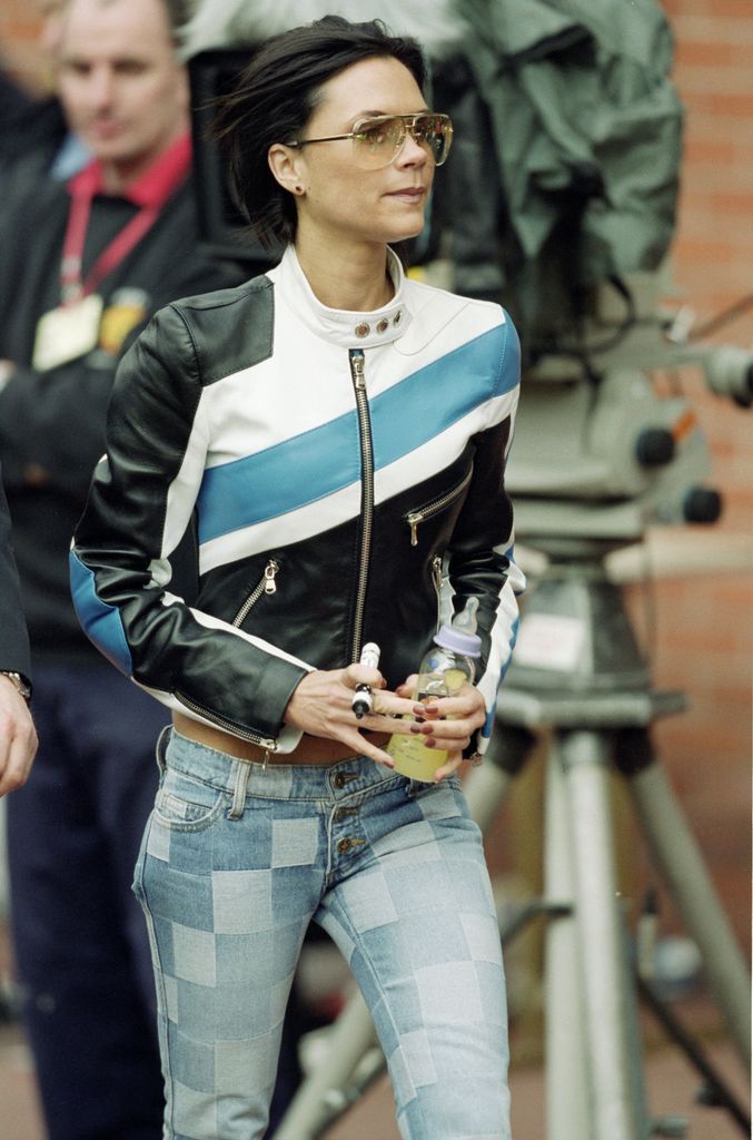 Victoria Beckham sporting a moto jacket, low slung jeans and yellow tinted square sunglasses in 2001. She carries a baby bottle as she is attending a Manchester United football game with her son Brooklyn Beckham