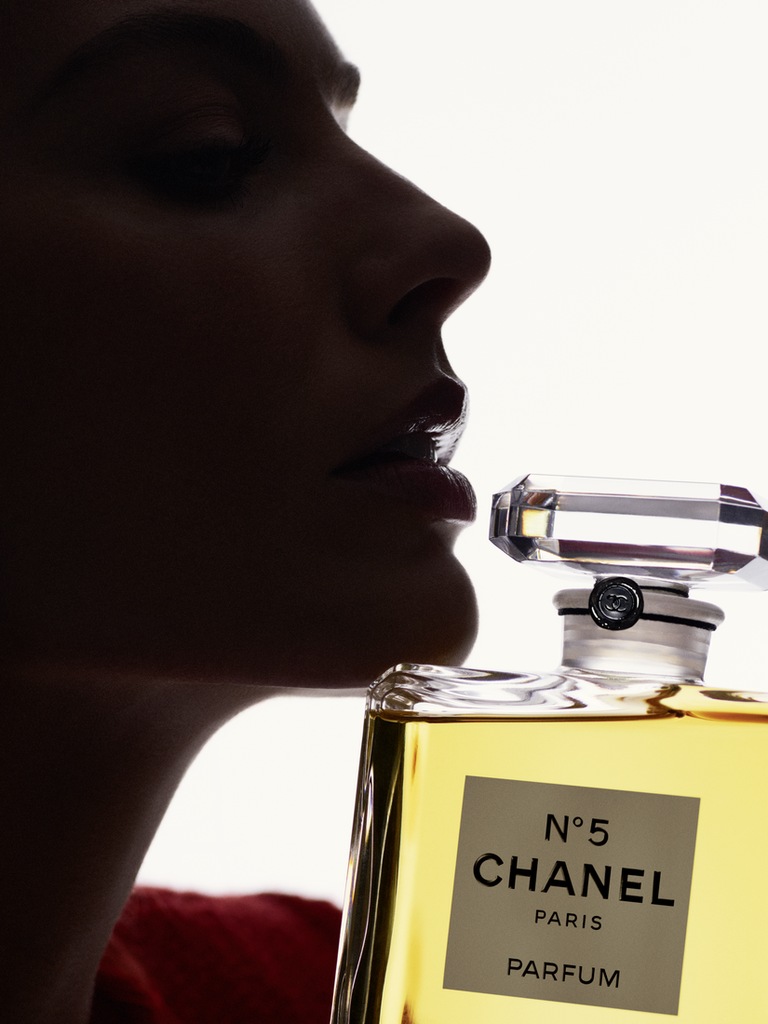 This image shows Margot Robbie in a close-up, silhouetted profile, emphasizing her facial features. She is positioned next to an iconic Chanel N°5 perfume bottle, with the bottle's top near her lips. The dramatic lighting creates a striking contrast, highlighting the luxury and timeless appeal of the fragrance.