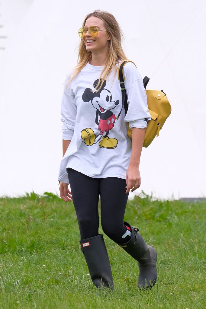 Margot Robbie attends day two of Glastonbury in 2017 wearing a white oversized sweatshirt with Mickey Mouse print, black leggings, Hunter wellies and yellow tinted sunglasses and yellow bag. 