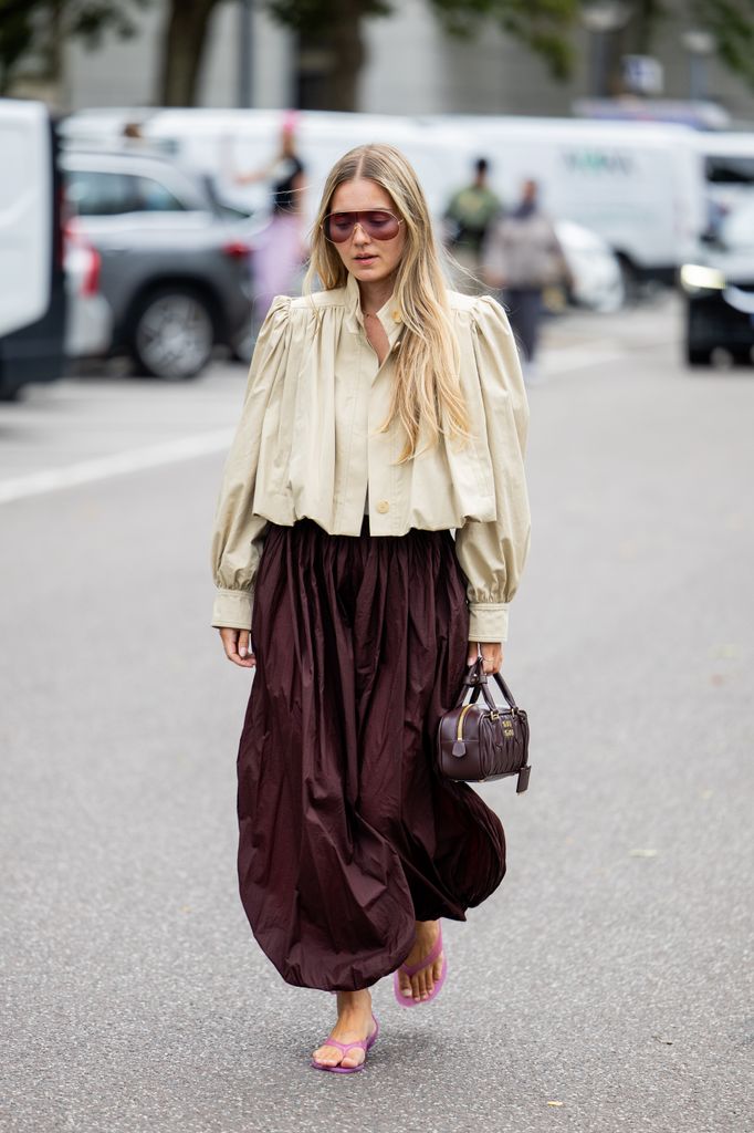 A guest wears wide leg burgundy skirt, beige jacket, Miu Miu bag, sunglasses outside MKDT Studio during Copenhagen Fashion Week day three