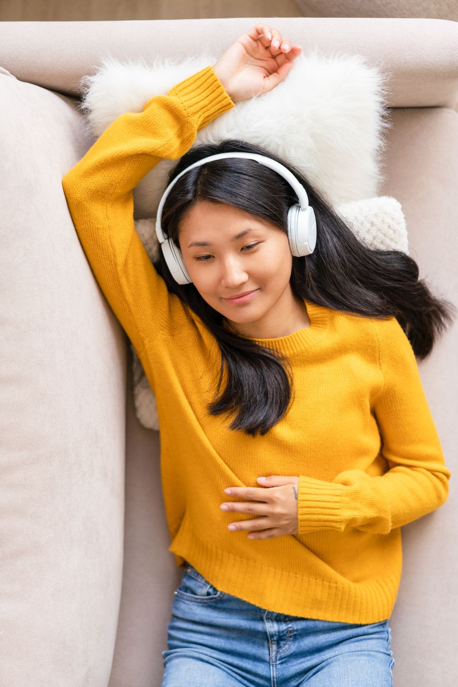 Happy asian woman lying on sofa listening to music in headphones. Enjoying audio playing in earphones. Relaxing, chilling at home. Positive emotions. Emotional spiritual health. Well-being, wellness with meditation. Enjoying good moments, slow life niksen
